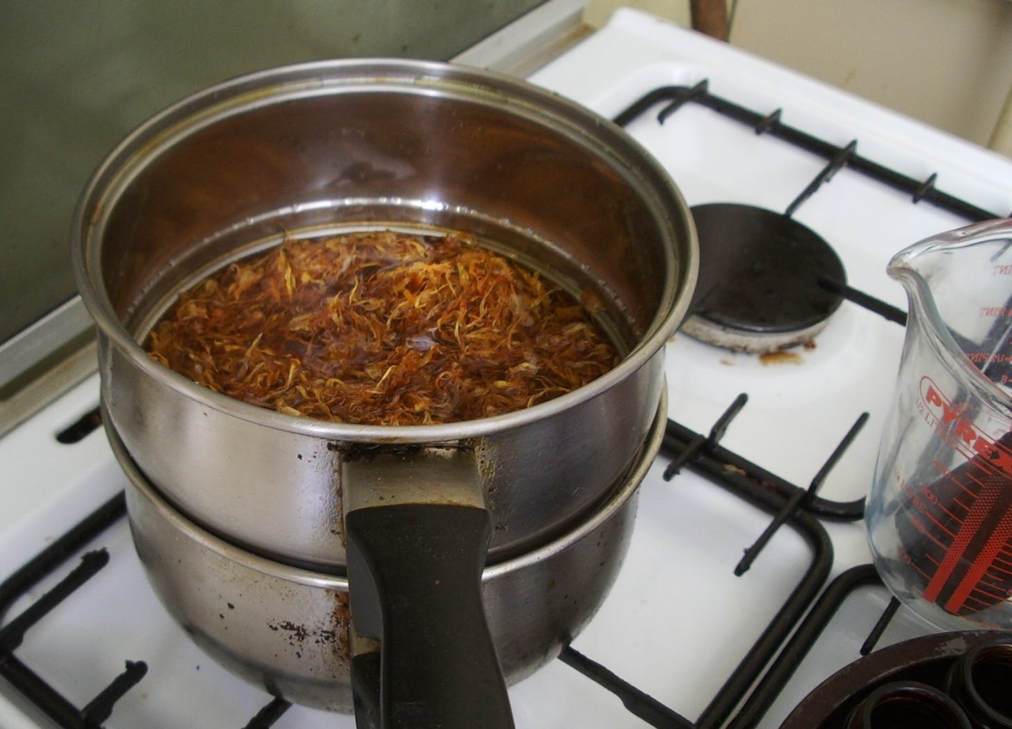 A double saucepan on a stove containing orange marigold petals in oil