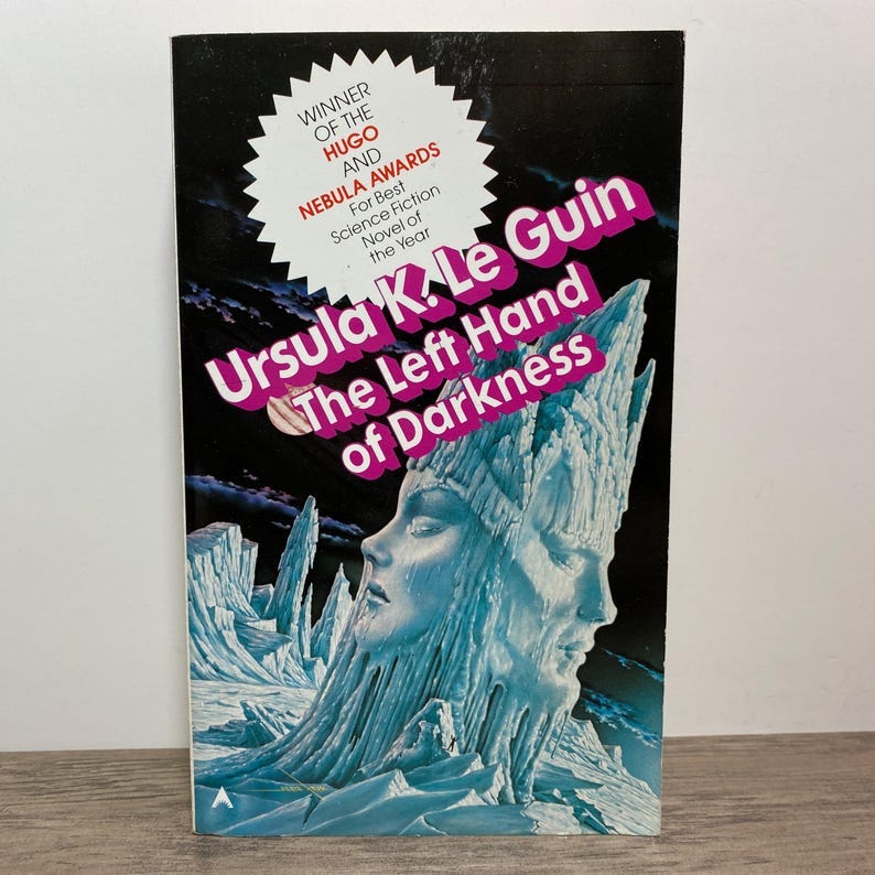May include: A book cover with a purple, white, and black color scheme. The title of the book is 'The Left Hand of Darkness' by Ursula K. Le Guin. The book cover features a landscape of ice and snow with a face carved into the ice. The book cover also includes a starburst with the text 'Winner of the Hugo and Nebula Awards'.