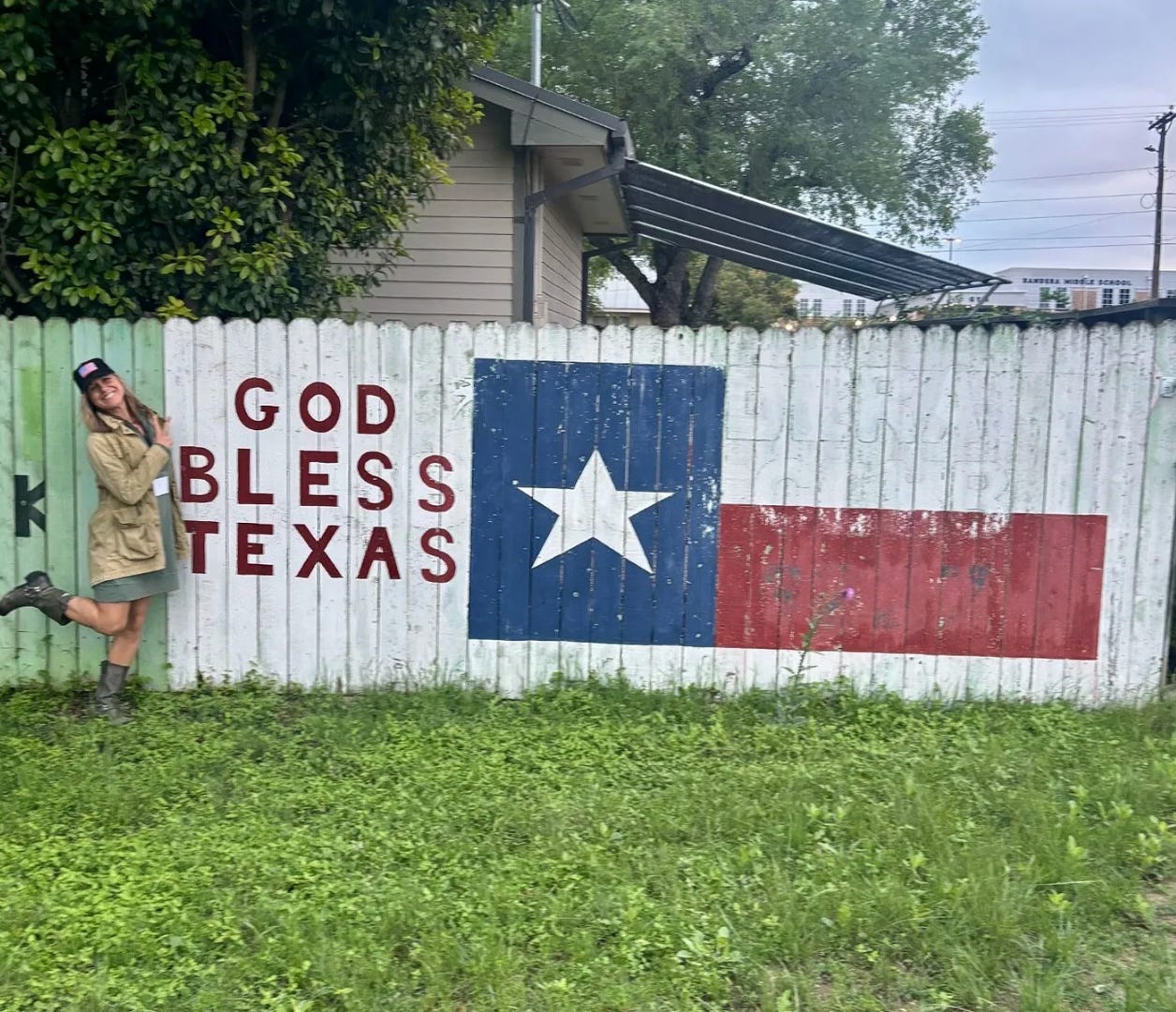 Woman poses playfully in front of a wooden fence painted with the Texas state flag and the words "God Bless Texas."