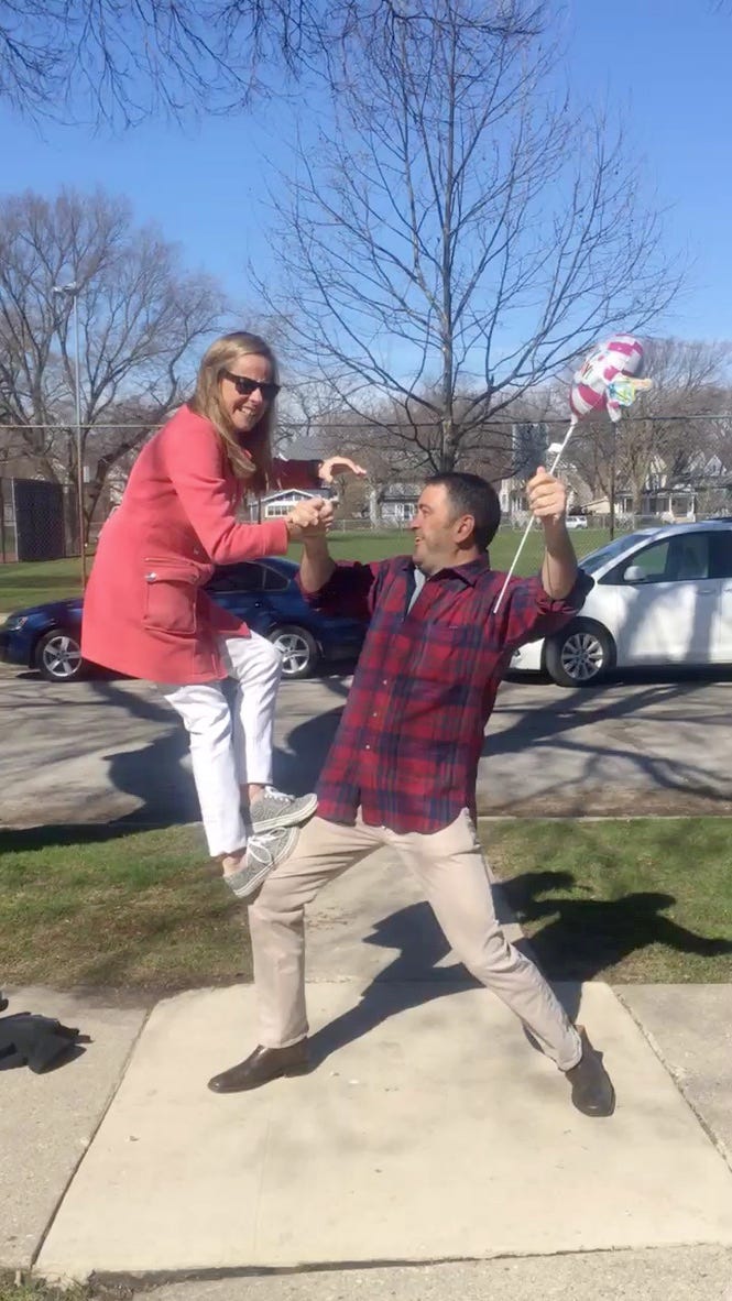 Blonde woman in sunglasses, salmon-colored jacket, white pants and gym shoes balances on the legs of a brown-haired man in a flannel shirt holding a birthday balloon. They are both smiling and being goofy. Blonde woman in sunglasses, salmon-colored jacket, white pants and gym shoes balances on the legs of a brown-haired man in a flannel shirt holding a birthday balloon. They are both smiling and being goofy.