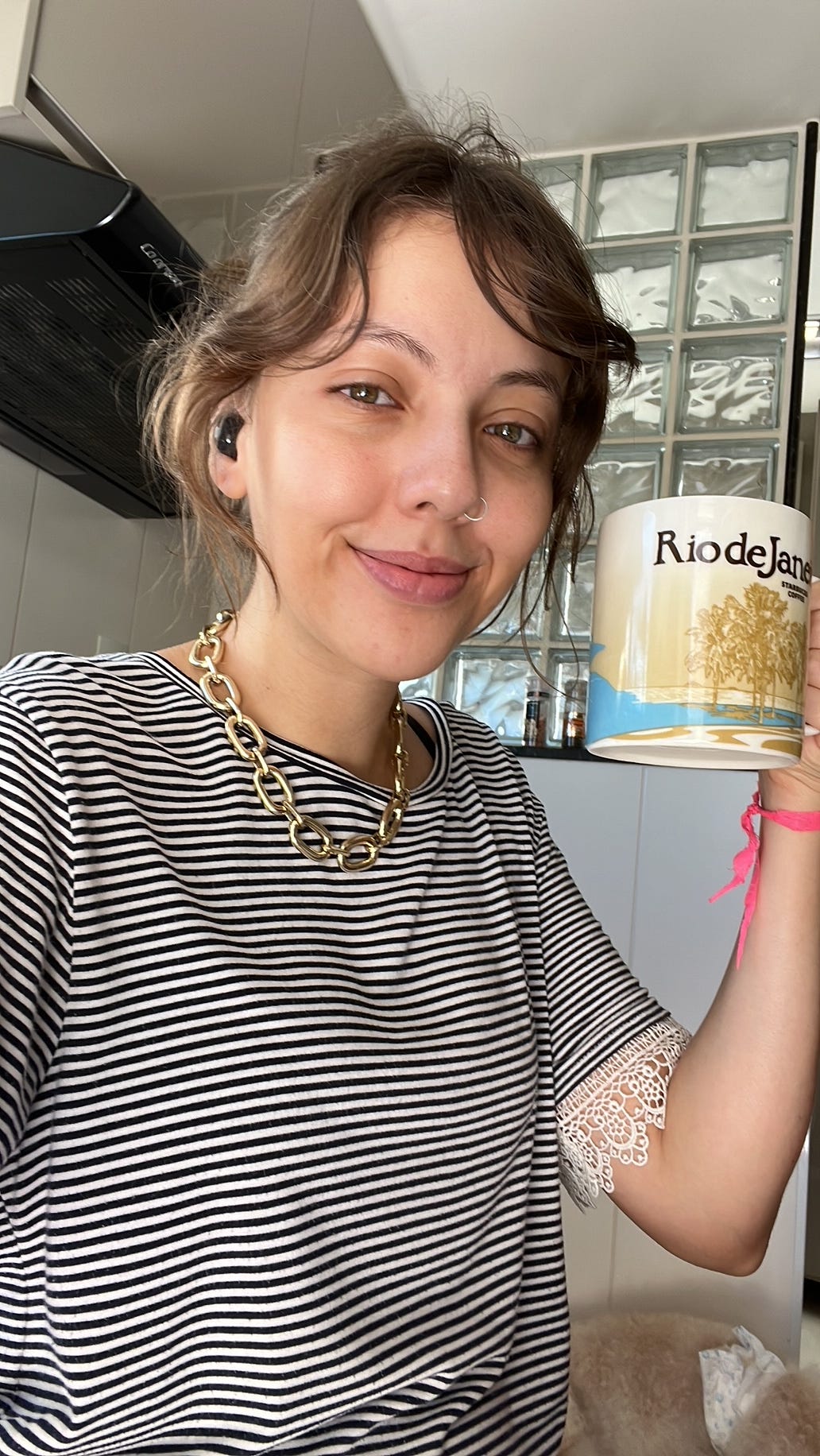 Selfie na cozinha com os olhos muito inchados. Estou de cabelo preso, blusa listrada, um sorrisinho, segurando uma enorme caneca temática do Rio de Janeiro