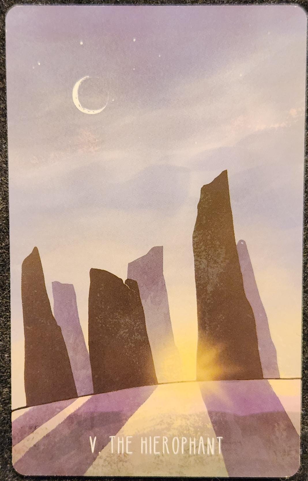A tarot card labelled V The Hierophant. The card shows a stone circle of large pillars. The sun is rising, peeking through the circle and casting long shadows on the ground. A crescent moon is visible in the light blue sky. 
