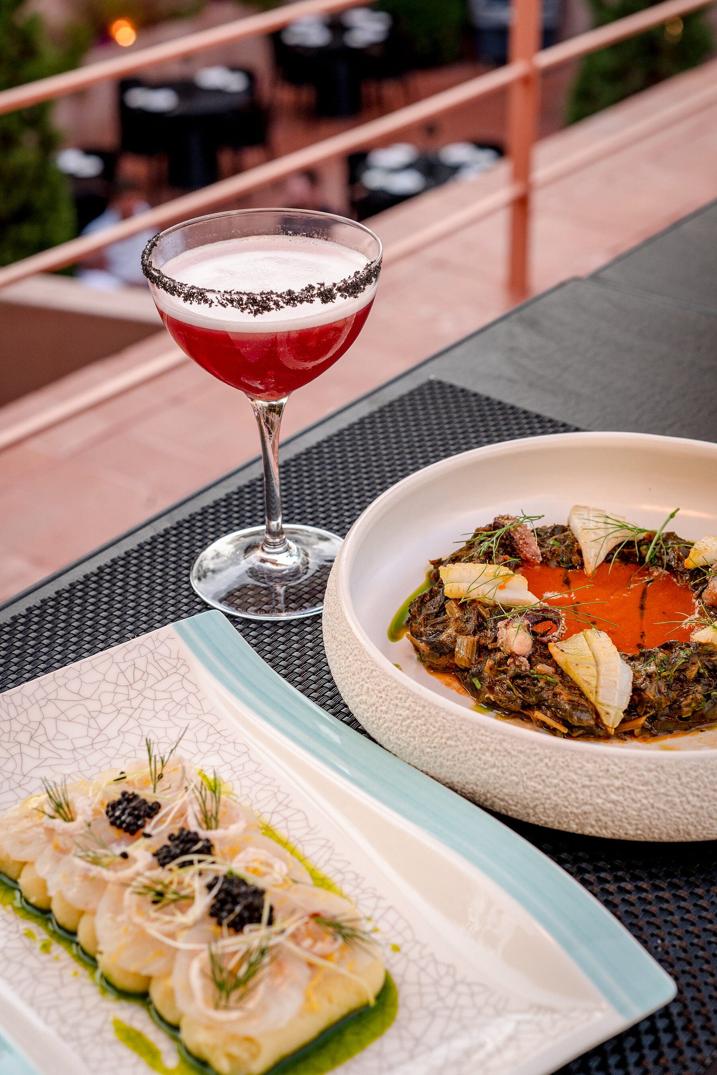 A table laden with refined dishes with delicate, fresh garnishes and a red-colored cocktail. 