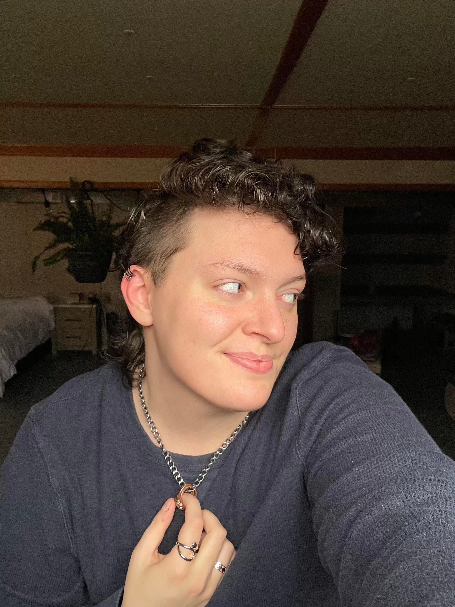 Alt-text: A person with short, curly brown hair and a side shave poses for a selfie, looking to the side with a soft smile. They wear a dark sweater, silver rings, and a chain necklace with a pendant, holding the necklace with one hand. The background features a cozy, dimly lit room with a bed, shelves, and a hanging plant.