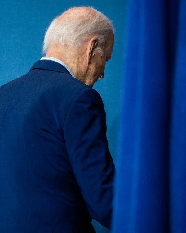 President Biden, with his back turned, walking past a blue curtain toward a blue background. President Biden, with his back turned, walking past a blue curtain toward a blue background.