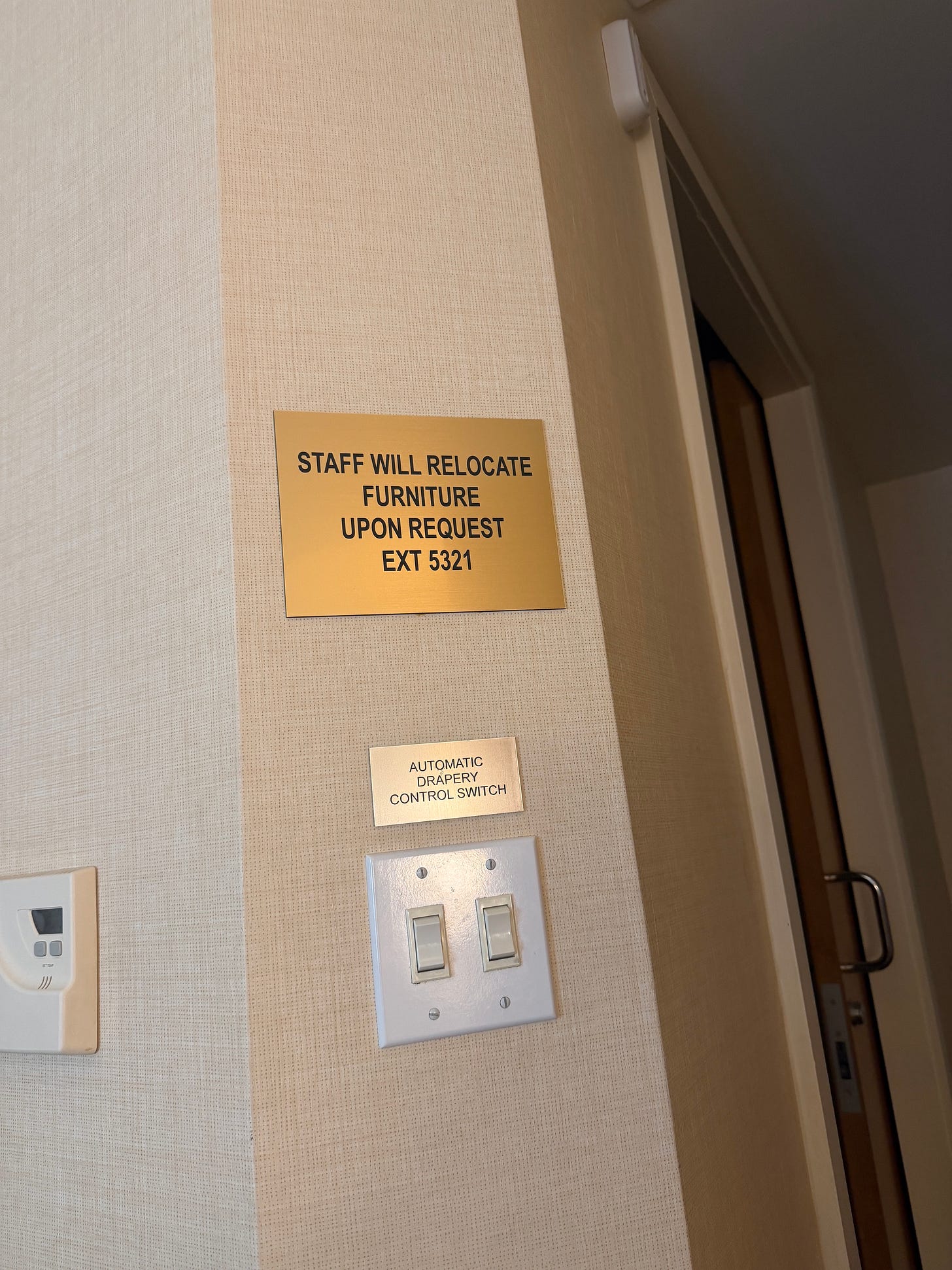 Image description: The corner of a beige wall, with two signs posted vertically, one on top of the other. The top is copper-colored, and says in black type, "Staff will relocate furniture upon request EXT 5321". Under it ias a smaller gold rectangle sign that says, "Automatic drapery control switch" above two cream-colored light switches.