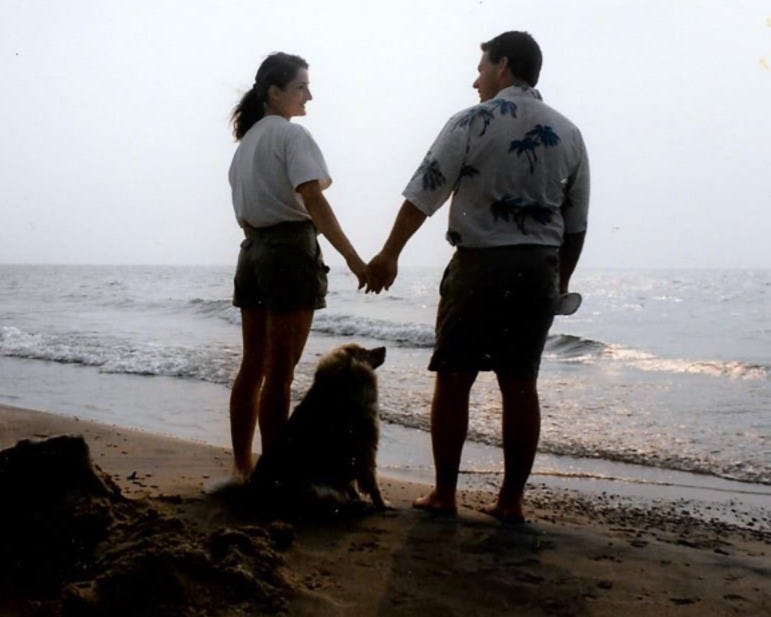 a young woman and man hold hands on a sandy beach with a dog between them