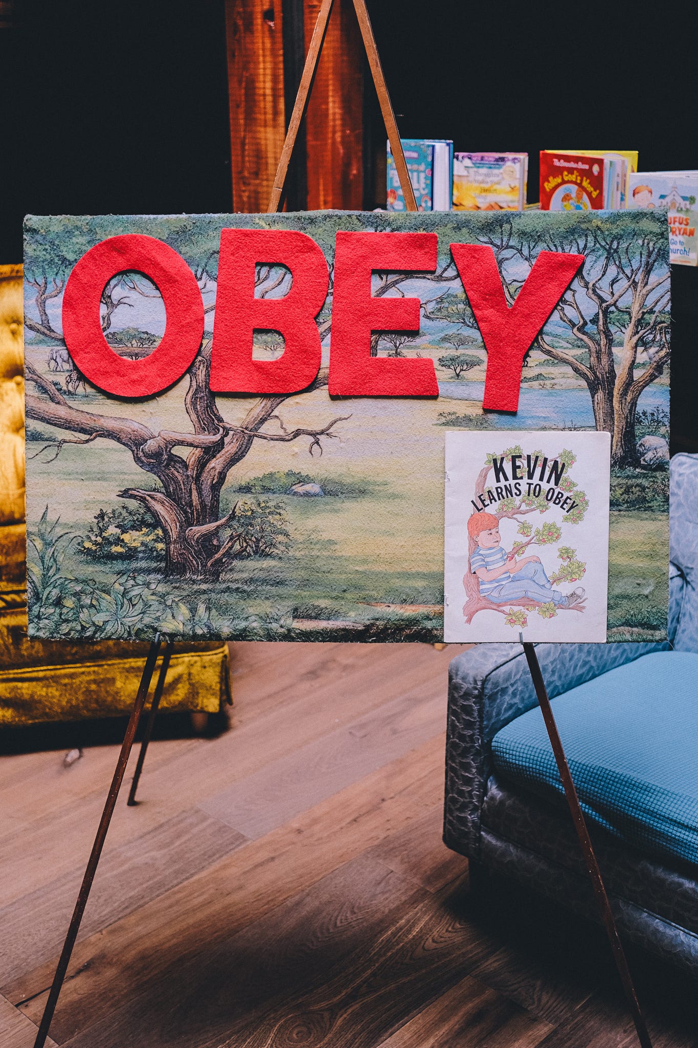 a felt board in a childern's area of a coffee shop with large red felt letters on it that say OBEY. there is also a book called kevin learns to obey on the board.