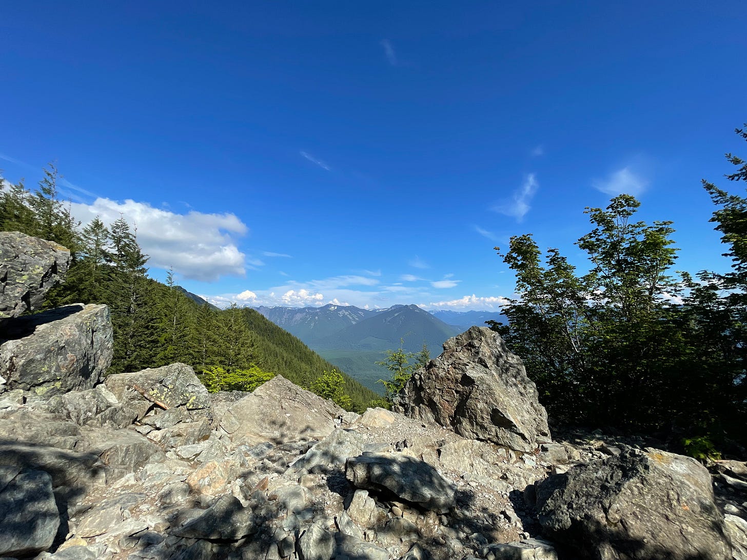 View from the top of Mt. Si, Washington. Distant blue mountain ranges, evergreen trees, and a rocky outcrop under a bright sunny sky. View from the top of Mt. Si, Washington. Distant blue mountain ranges, evergreen trees, and a rocky outcrop under a bright sunny sky.