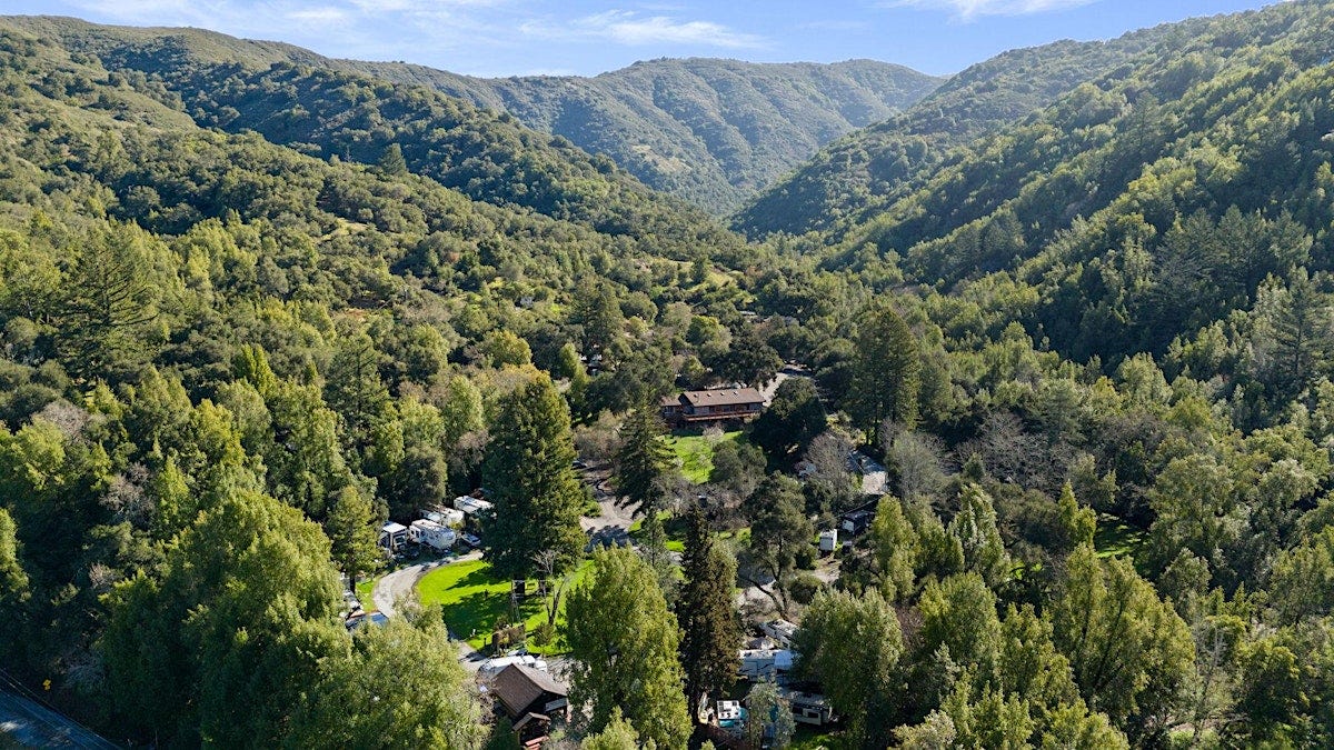 Aerial view of Lupin Lodge nestled in the Santa Cruz Mountains, surrounded by dense green forest and rolling hills, with cabins, RVs, and open spaces visible among the trees.