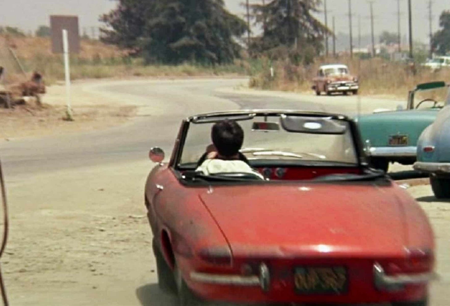 Actor drives red Alfa Romeo Spider convertible away from the camera across a dusty petrol station forecourt towards the curving Californian highway. The car is battered and dirty.