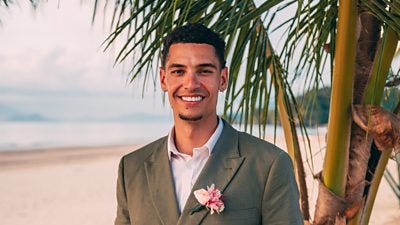 A man in a grey suit jacket and light shirt smiles as he stands on a beach