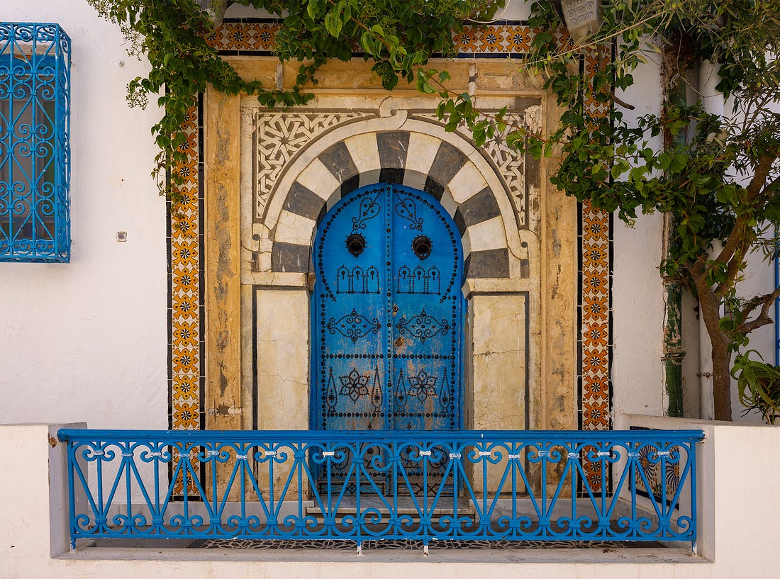 Traditional blue door in Sidi Bou Said framed by an arched limestone and marble doorway, patterned tiles, and cascading vines. The intricate ironwork and symmetry reflect the village’s Andalusian-Maghrebi heritage and its enduring balance between ornament, function, and light. Traditional blue door in Sidi Bou Said framed by an arched limestone and marble doorway, patterned tiles, and cascading vines. The intricate ironwork and symmetry reflect the village’s Andalusian-Maghrebi heritage and its enduring balance between ornament, function, and light.