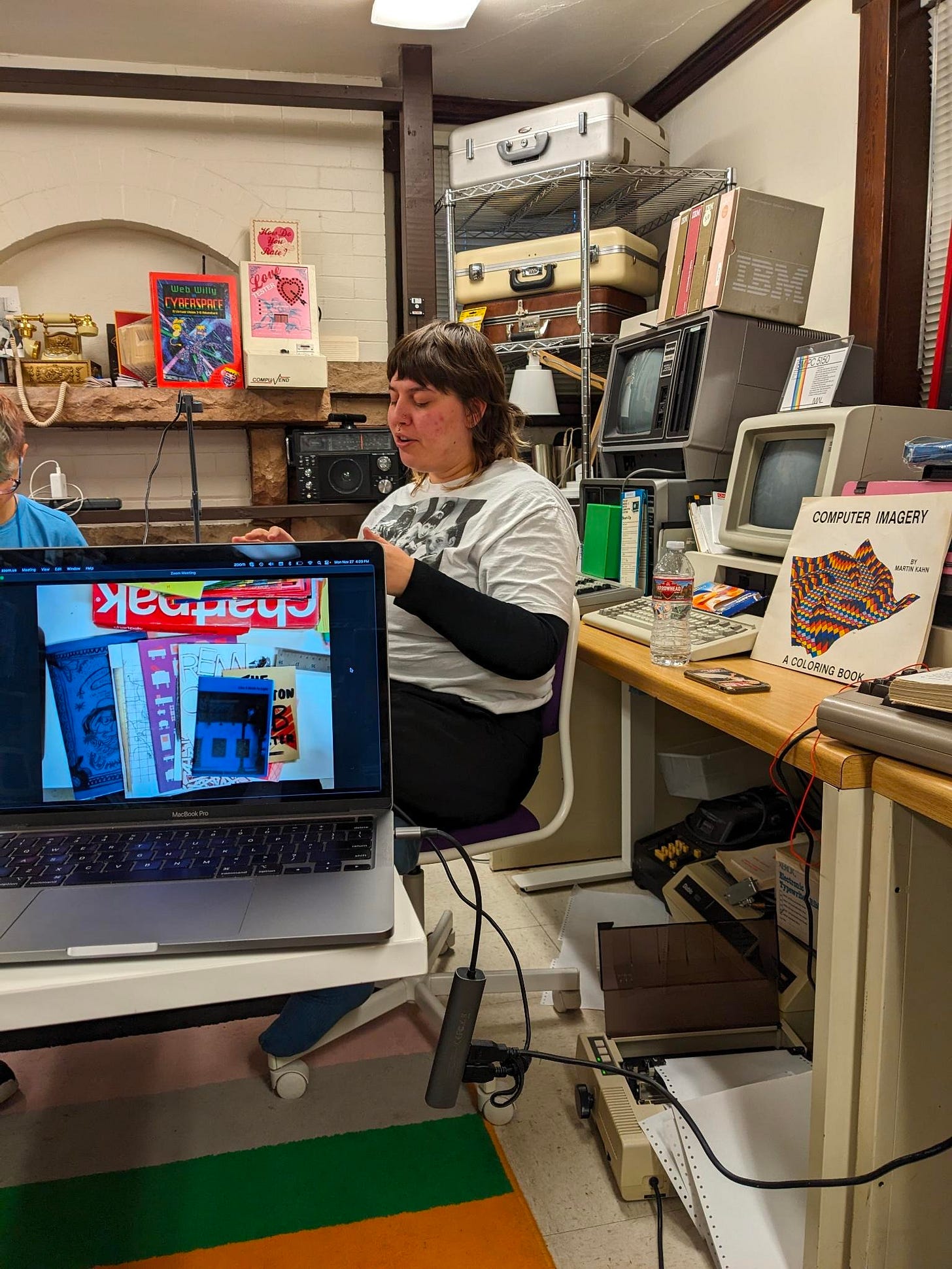 Sid Drmay sits at a table full of craft supplies, explaining zinemaking as an artistic practice. A computer in the front shows a camera shot of the example zines Sid is sharing, 