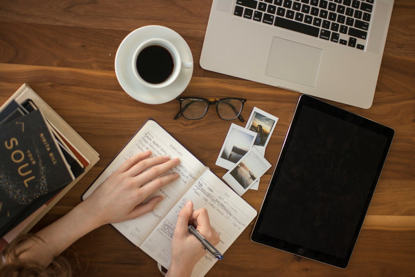Overhead view of a wooden desk with a laptop, a cup of black coffee, a tablet and a hand writing in a paper notebook. Overhead view of a wooden desk with a laptop, a cup of black coffee, a tablet and a hand writing in a paper notebook.
