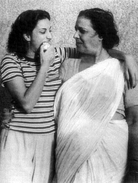 A picture of young Nargis munching on an apple in a striped tshirt and trousers with one arm around her mum, Jaddan Bai, who is dressed in a white saree and blouse. The picture is black and white. The two are looking fondly at each other.