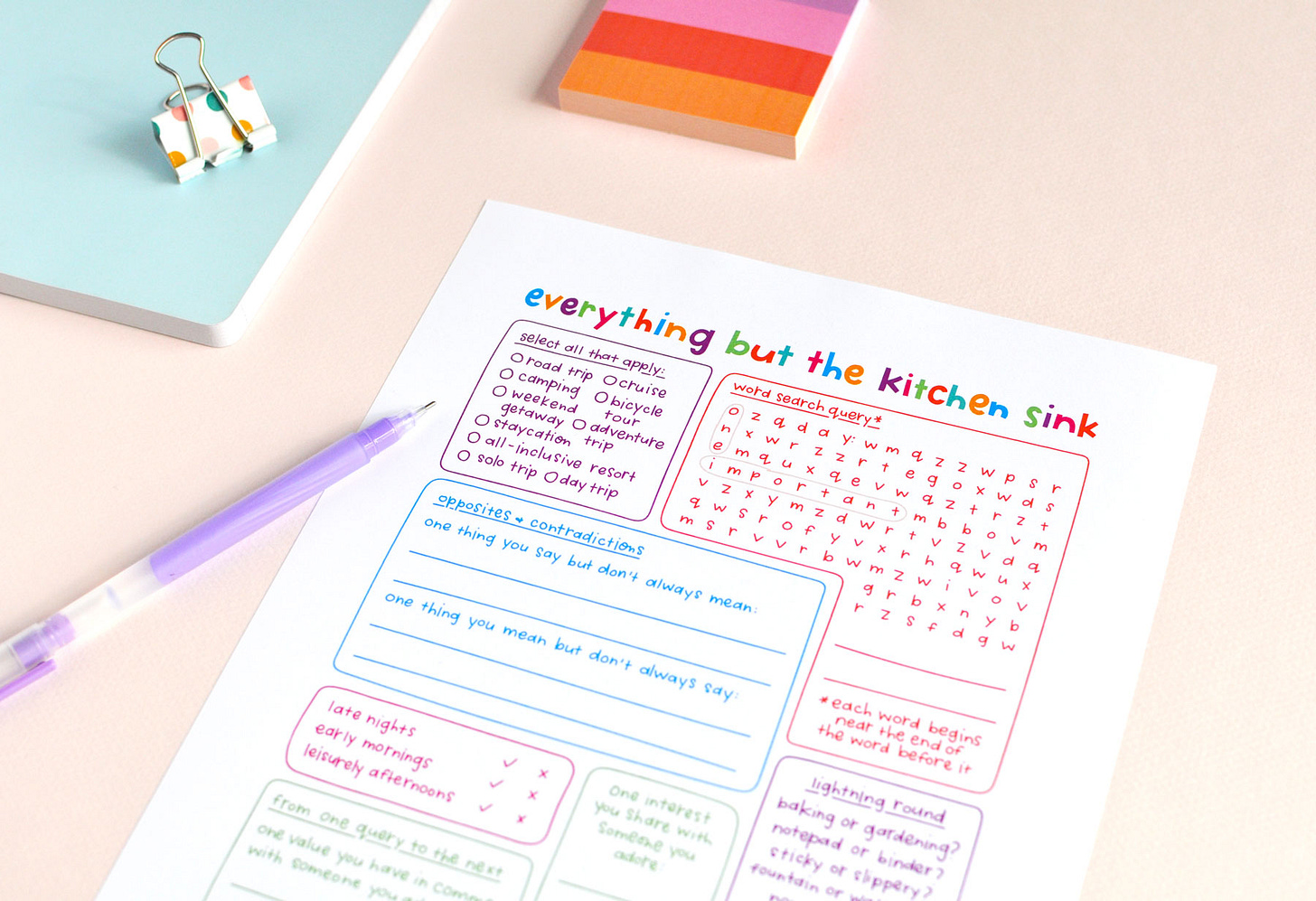 A colourful, hand-lettered journal page titled Everything But the Kitchen Sink filled with prompts and puzzles to inspire your journaling. Shown with colourful notepad, notebook, binder clip and purple pen.