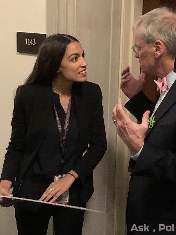 AOC holds a signs as she talks to a man in a suit with a green bicycle pin on his suit