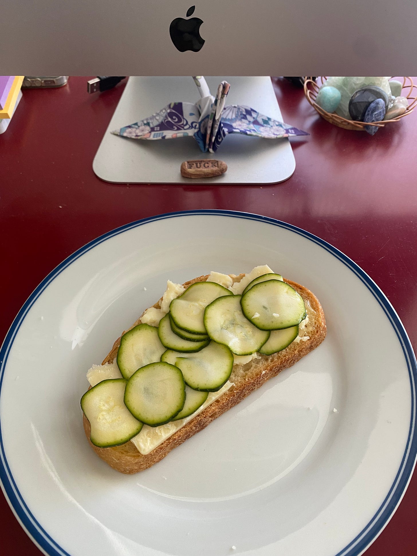 On my desk in front of my computer, a white plate with a blue rim holding an open-faced cheese and zucchini pickle sandwich on sourdough.