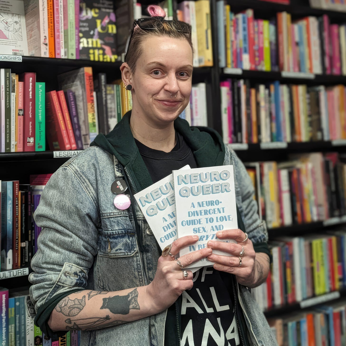 CJ DeBarra standing in a bookshop, holding two copies of Neuroqueer: A Neurodivergent Guide to Love, Sex, and Everything in Between. They wear a denim jacket over a dark T-shirt, with short hair, visible tattoos, and enamel pins on their jacket. Bookshelves filled with brightly coloured spines line the background.
