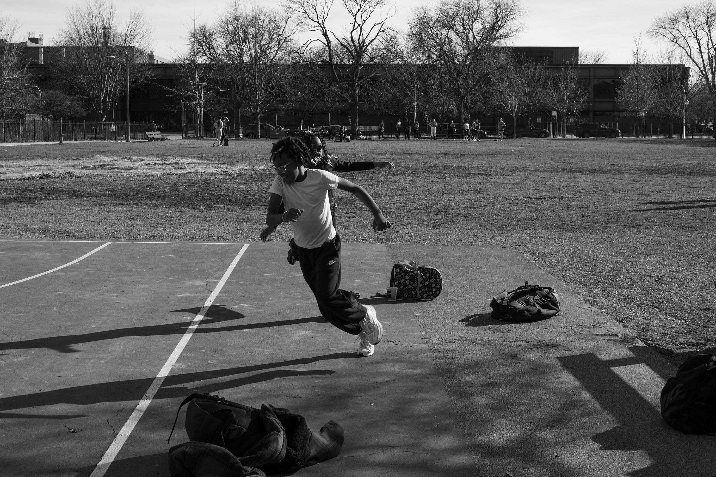 A black-and-white photograph of two young kids sprinting across the edge of a basketball court, their bodies leaning forward in motion. One boy in the foreground wears glasses and a white t-shirt, his arms swinging as he runs, while the other kid follows closely behind. Shadows stretch across the pavement, adding depth and movement to the scene. Scattered backpacks and belongings lie on the ground, while in the background, a park with trees, people, and an urban school building sets the stage for this moment of youthful energy and play.