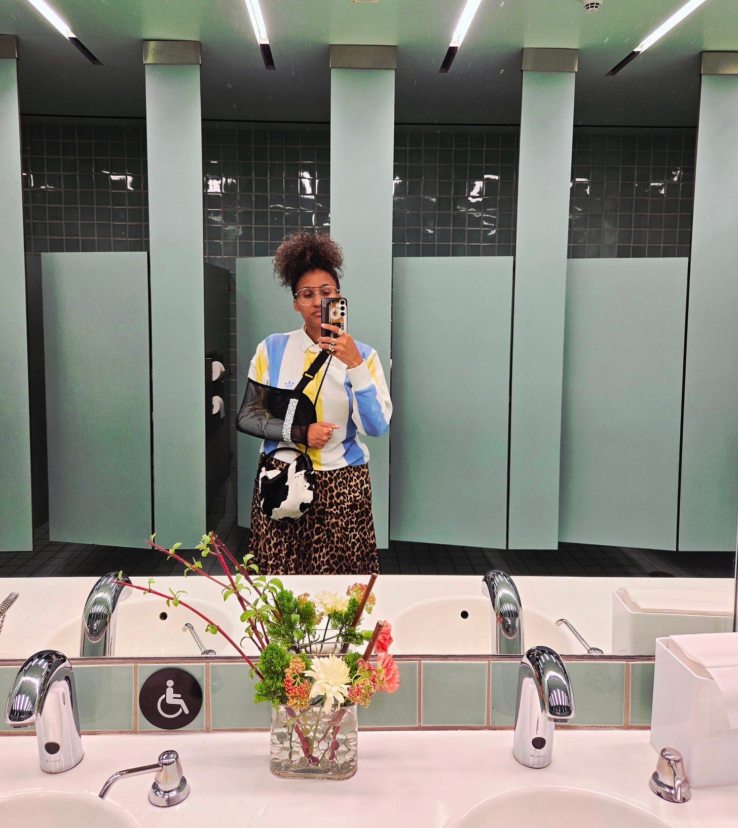 Dr. Cadet takes a mirror selfie in a seafoam green bathroom, like ties, ceiling, doors, seafoam green. She wears a vintage Adidas long sleeve blue, white and yellow striped polo with a leopard print skirt and her cross body emotion support Freda Salvador cowhide purse. With her curly hair gathered on top of her head her black mesh sling adorned with pearls by Dr. Cadet is the star of the show.
