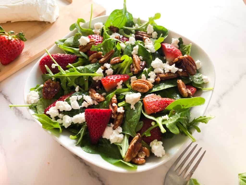 strawberry goat cheese salad in a white bowl with candied pecans and arugula and baby spinach with a bowl of strawberries