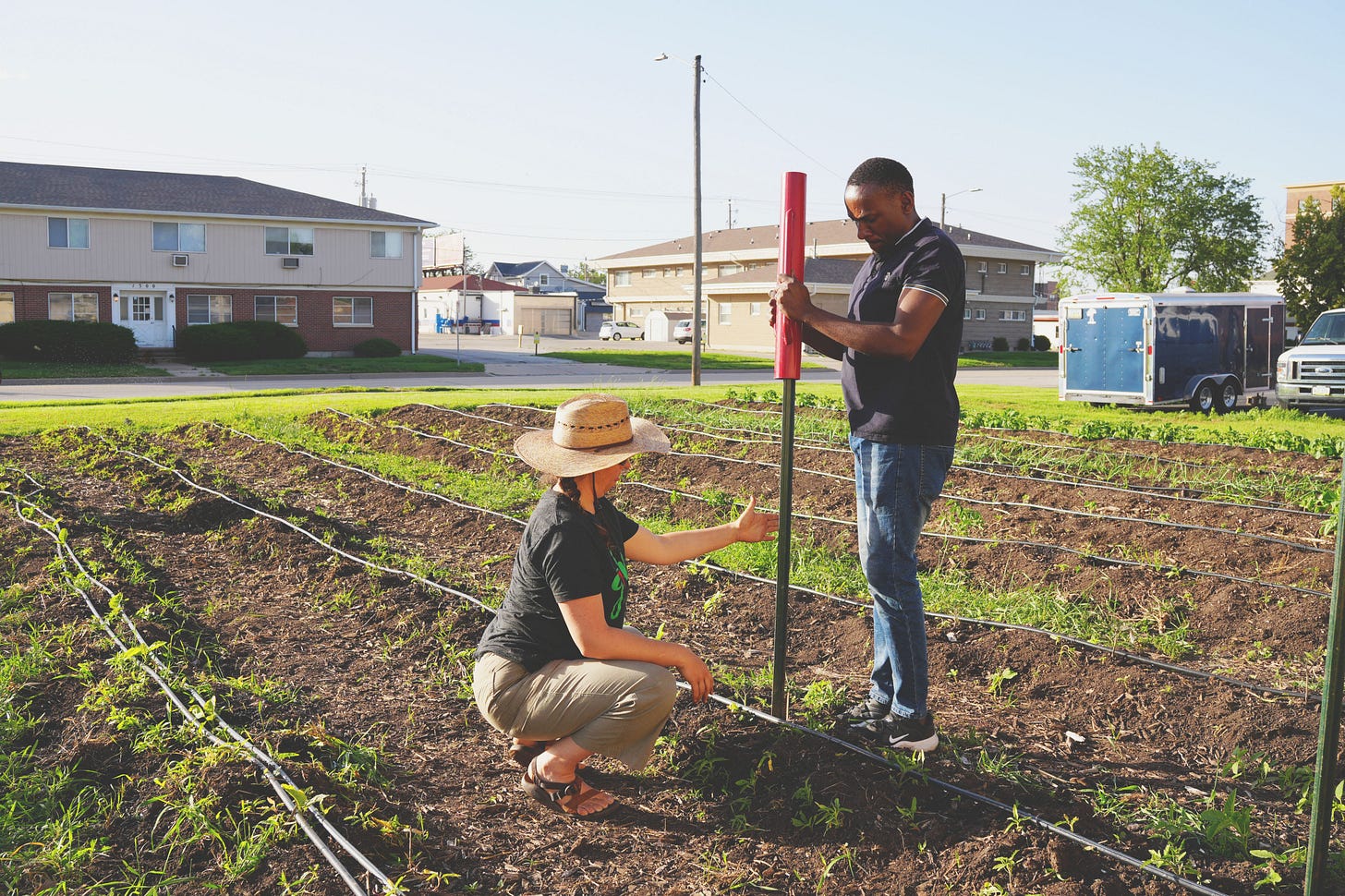 A woman in a sun hat crouches to help make sure a stake is properly put into the ground as a man stands using a tool to pound the stake into the soil of a garden plot they are standing in the middle of. A woman in a sun hat crouches to help make sure a stake is properly put into the ground as a man stands using a tool to pound the stake into the soil of a garden plot they are standing in the middle of.