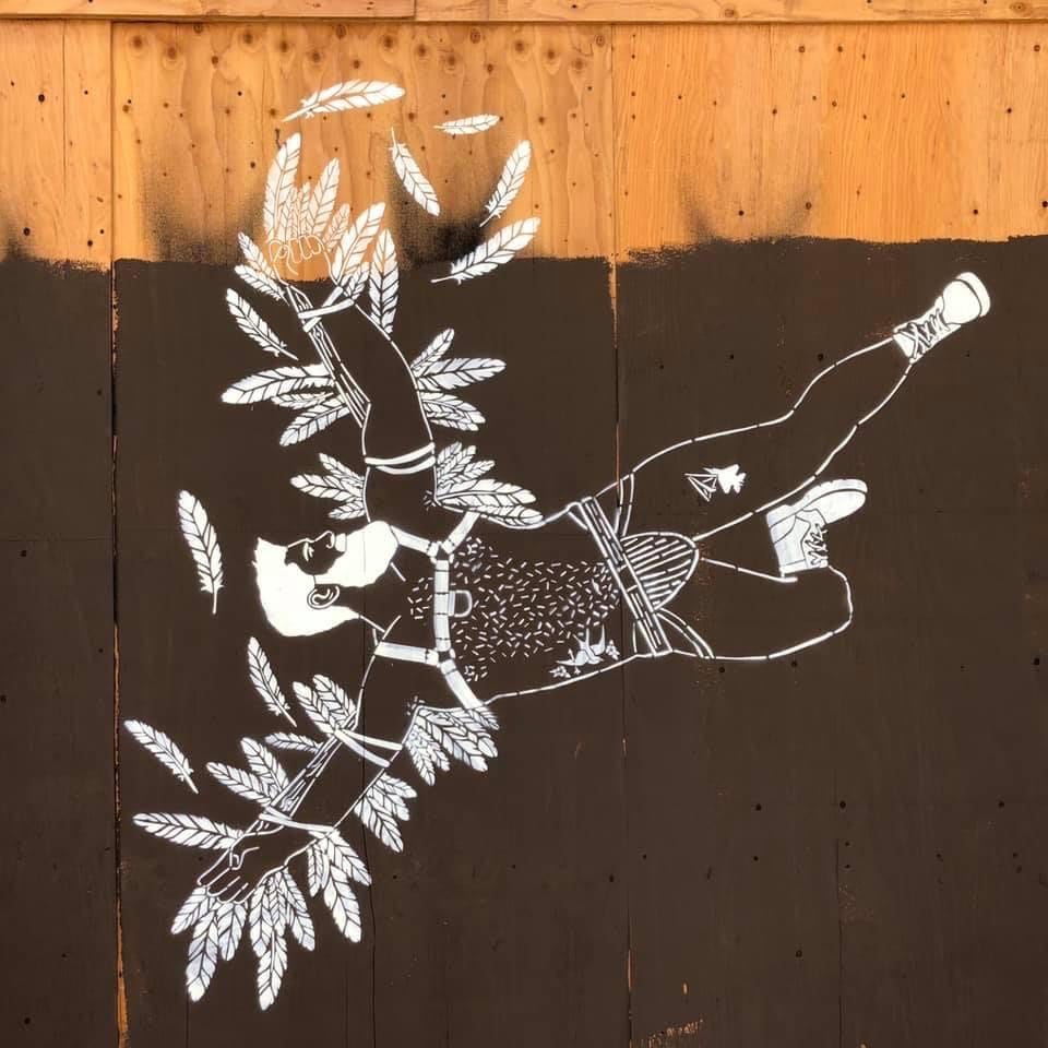 Mural of a nude man in a chest harness and jock strap surrounded by white feathers on a wooden wall. The figure appears to float peacefully, evoking a sense of freedom. He is wearing white boots, and his arms are out by his sides. The feathers are tied to his arms with rope. He has a white beard and hair, and his head is turned to the side. He has a hairy chest. The board he is on is mostly painted black, with the original wood color showing on top. Mural of a nude man in a chest harness and jock strap surrounded by white feathers on a wooden wall. The figure appears to float peacefully, evoking a sense of freedom. He is wearing white boots, and his arms are out by his sides. The feathers are tied to his arms with rope. He has a white beard and hair, and his head is turned to the side. He has a hairy chest. The board he is on is mostly painted black, with the original wood color showing on top.