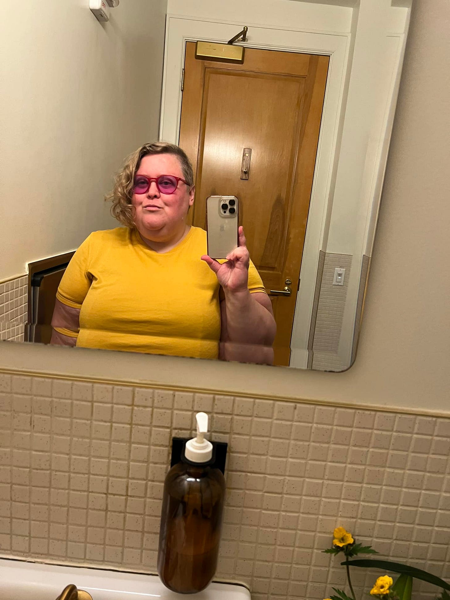 Alison, a fat white woman with blonde asymmetrical hair wearing red glasses indoors, in other words me. I’m wearing a yellow short sleeve top.
