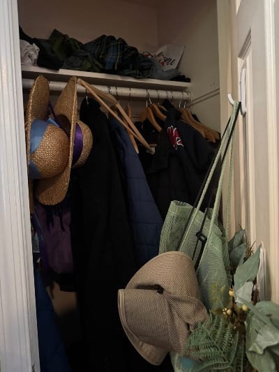 Small closet with door propped open. Seen are a pile of random items on top, items hanging from the door, and general disarray