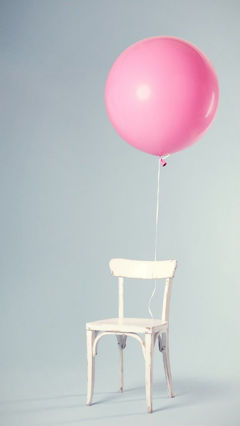 Minimalist image of a white chair with a pink balloon tied to the back, set against a soft gray background.