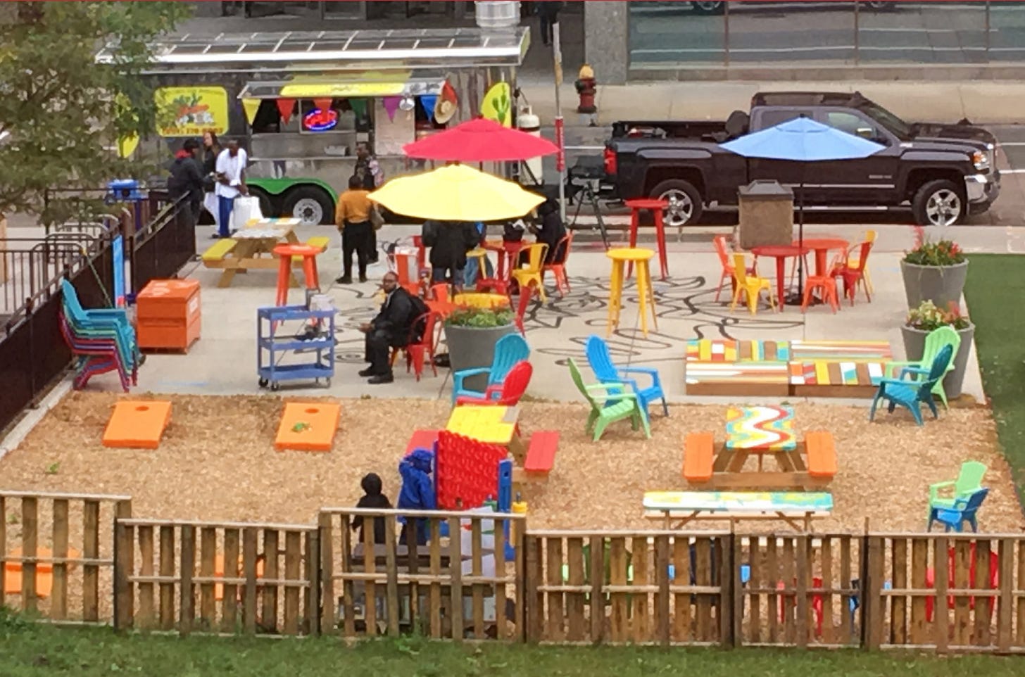 This photo shows Central Station with major improvements. There is a fun-looking fence, a food truck, outdoor games, and colorful shaded tables and chairs.