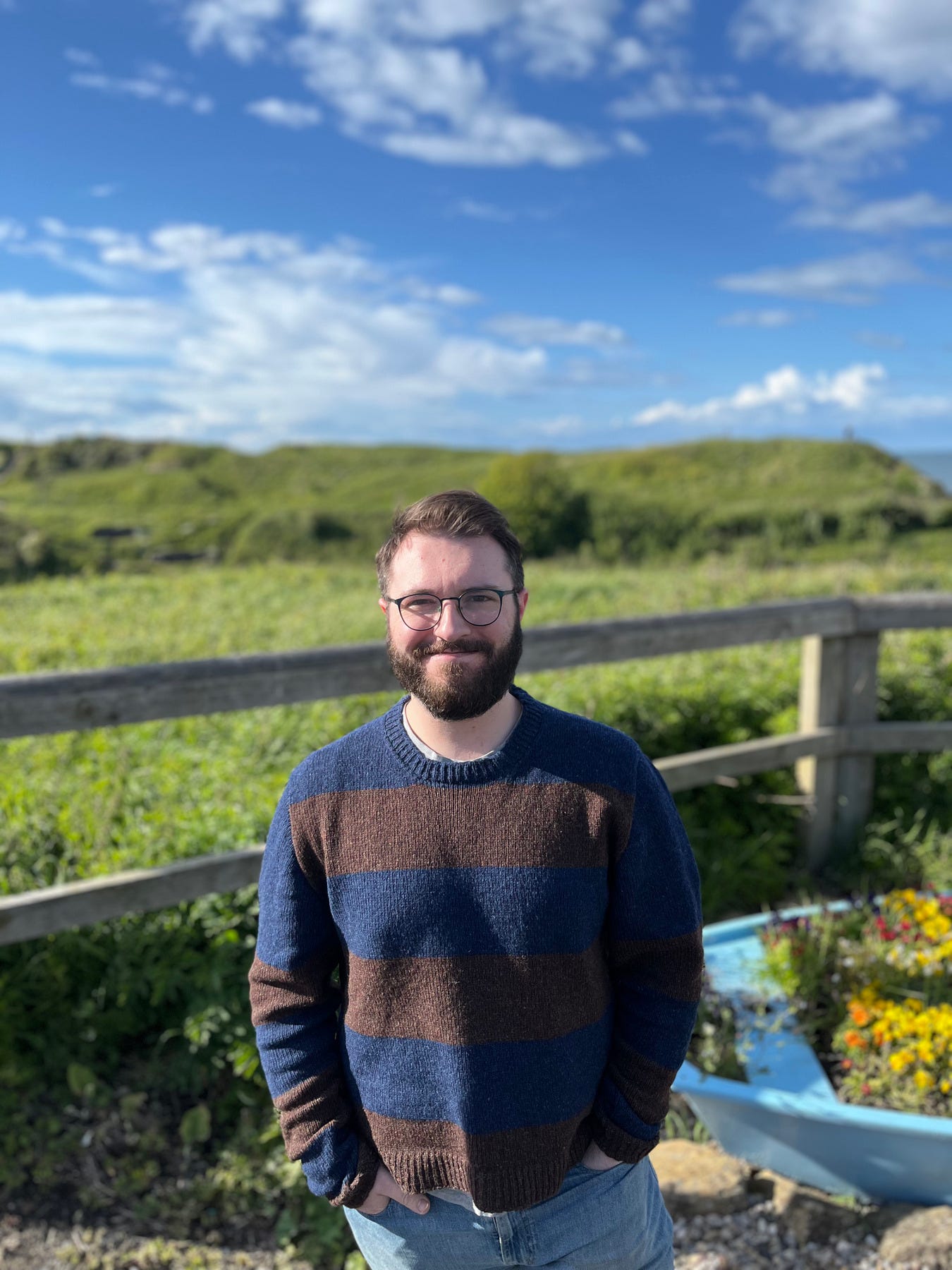 A man wearing a wide-stripe chocolate and navy sweater standing outdoors at Seaton Sluice, smiling, green coastal headland and blue sky behind him.