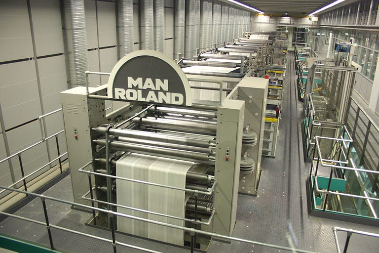 An offset press. It is a massive industrial machine dozens of feet long, from start to finish, and so tall it needs a multi-level scaffold for workers to navigate.