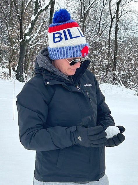 Woman in a Bills beanie and black jacket holds a small snowball, and then a basketball-sized snowball, and then stands next to a giant snowball that is almost as tall as her.