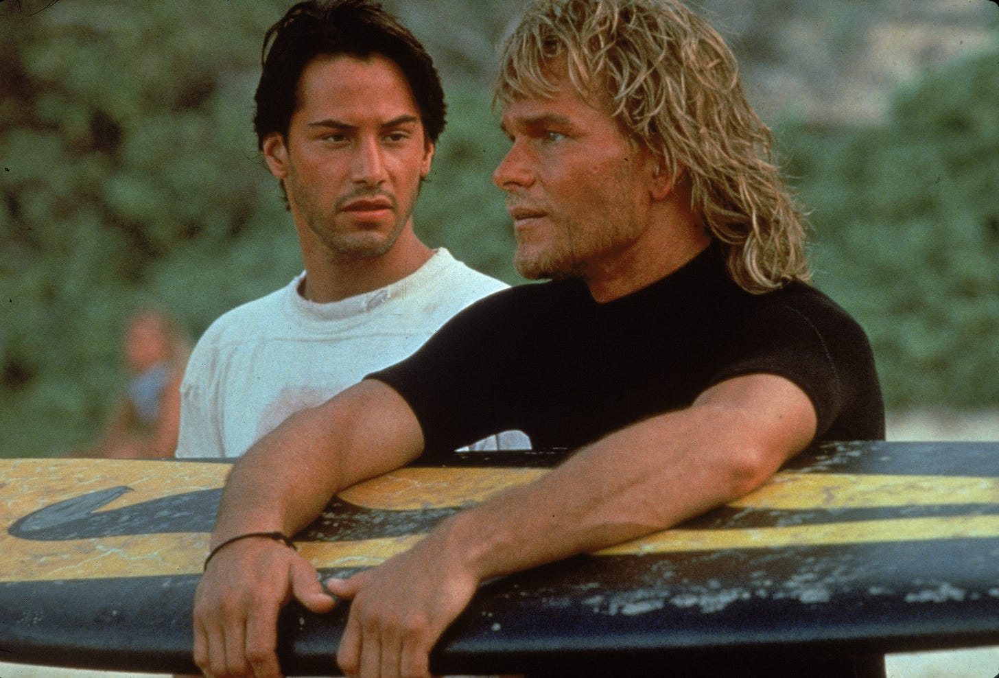 A picture from Point Break. Keanu Reeves looks at Patrick Swayze holding a surf board. They are both very hot men.