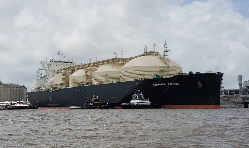 Photo of huge liquefied natural gas (LNG) tanker, with four big domed gas storage tanks, being aided by tugboats in port.