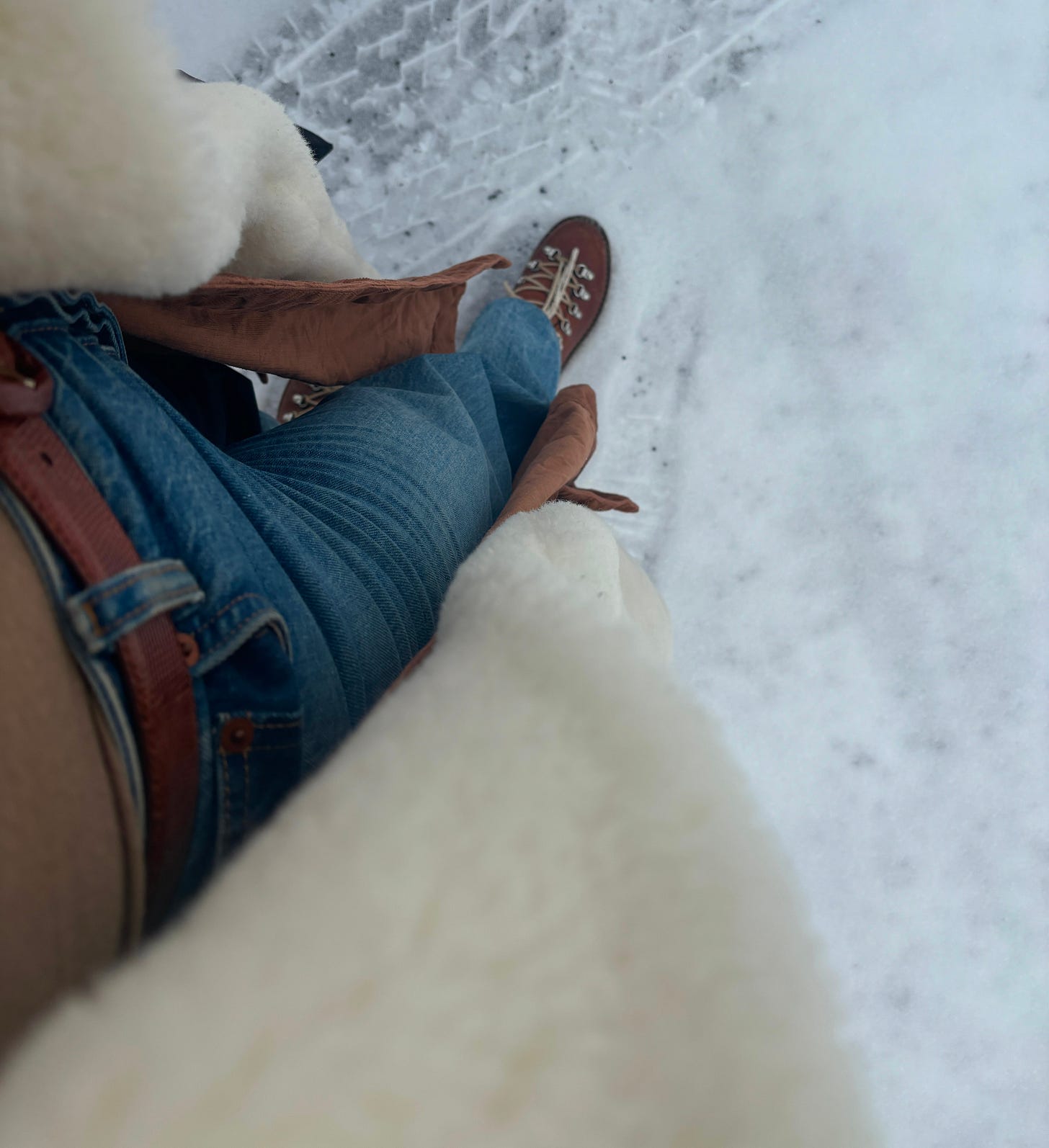 a birdseye shot of an outfit from the perspective of the wearer. it shows fitting jeans, a brown belt, and open brown shirt dress, and a wool overcoat. italian brown leather boots step in the snow, which covers the ground.