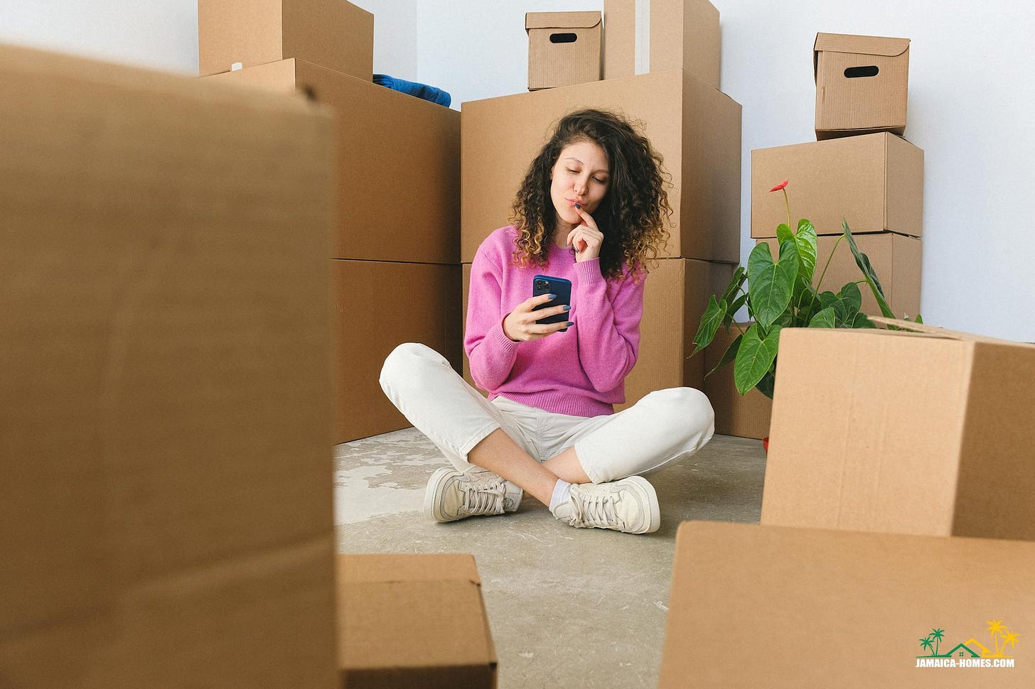 excited young woman messaging on smartphone sitting on floor near boxes after relocation