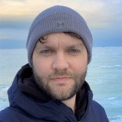 Um homem com barba curta, pele branca, olhos azuis e expressão neutra. Usa touca cinza e um casaco azul-escuro com zíper. Ao fundo, há uma paisagem de uma superfície de gelo que se estende até o horizonte e o céu parcialmente nublado.