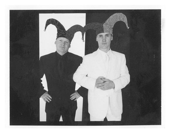 Two men (Tom and David Gardner), one in an all-black suit and the other in an all-white suit, both wearing jester caps, in front of black and white backgrounds. Two men (Tom and David Gardner), one in an all-black suit and the other in an all-white suit, both wearing jester caps, in front of black and white backgrounds.