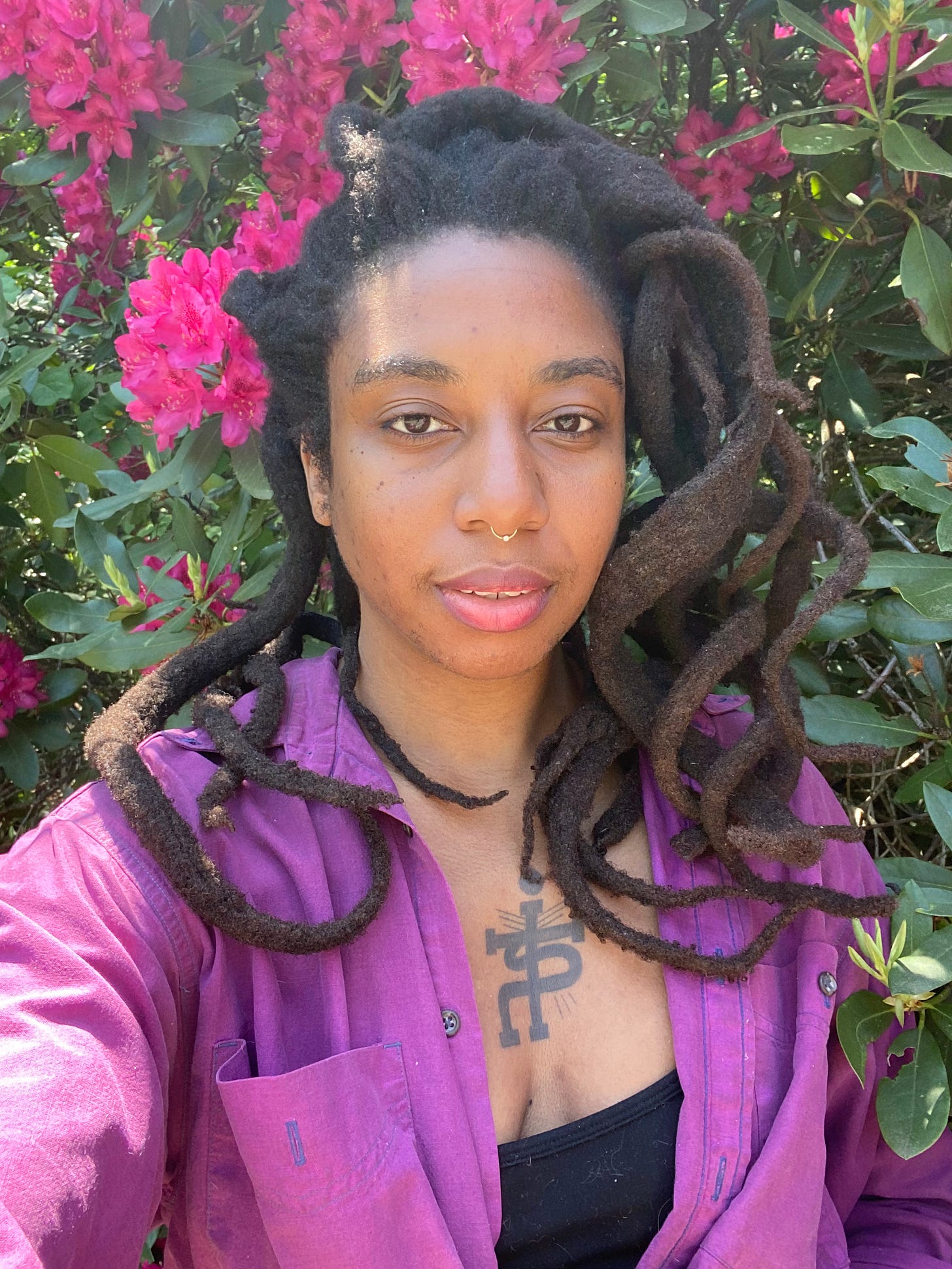 beautiful black woman with long, loosely curled freeform locs. She is wearing a purple shirt and black tank top with a large symbol tattoo on her chest. She is standing in front of a large bush with green leaves and pink flowers.