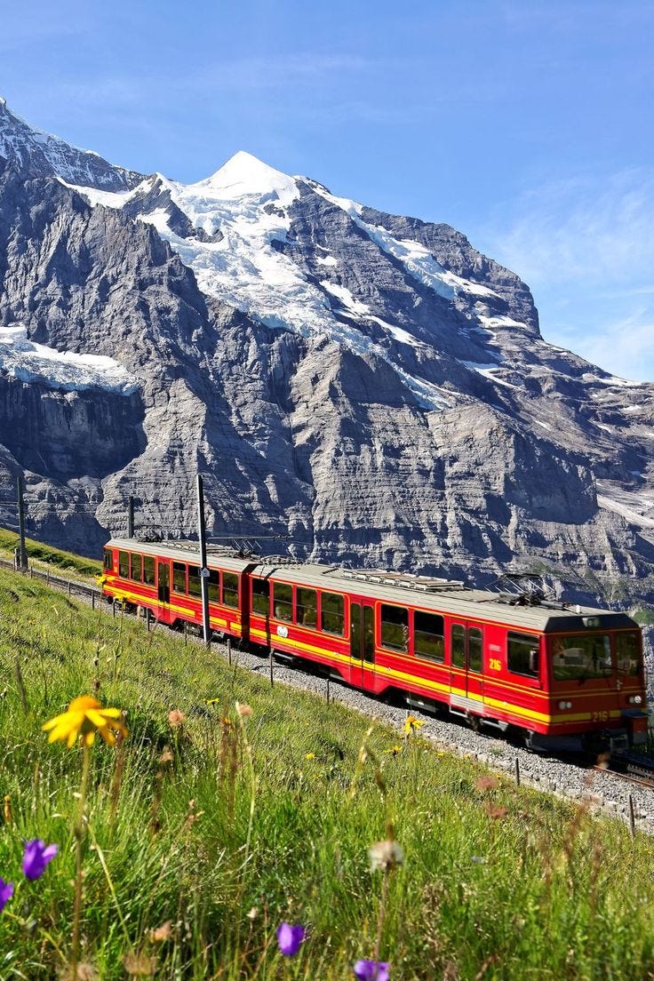 This may contain: a red and yellow train traveling through a lush green hillside covered in snow capped mountains
