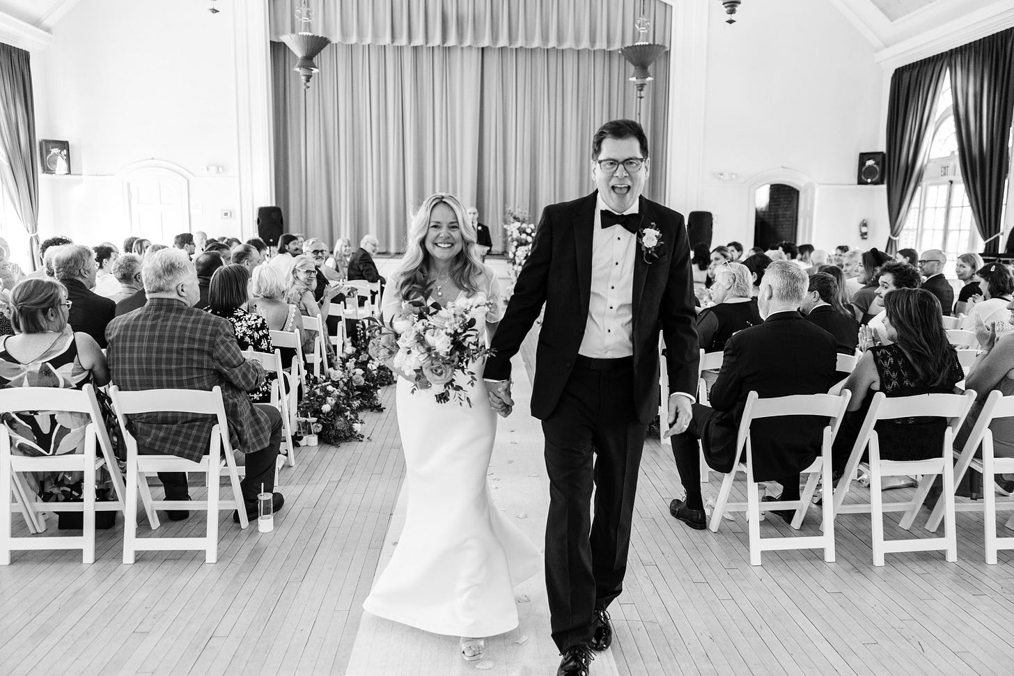 Bride and groom smile for camera. Bride is holding flowers. Groom wears a tuxedo and glasses. Wedding guests sit in white folding chairs. A curtained stage is behind them. 