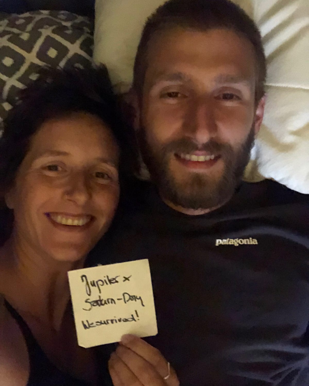 Two smiling people lying side by side on a bed, one holding a handwritten sign that says "Jupiter × Saturn-Day — We survived!" The person on the right wears a black Patagonia shirt, and the mood appears joyful and relieved. Two smiling people lying side by side on a bed, one holding a handwritten sign that says "Jupiter × Saturn-Day — We survived!" The person on the right wears a black Patagonia shirt, and the mood appears joyful and relieved.