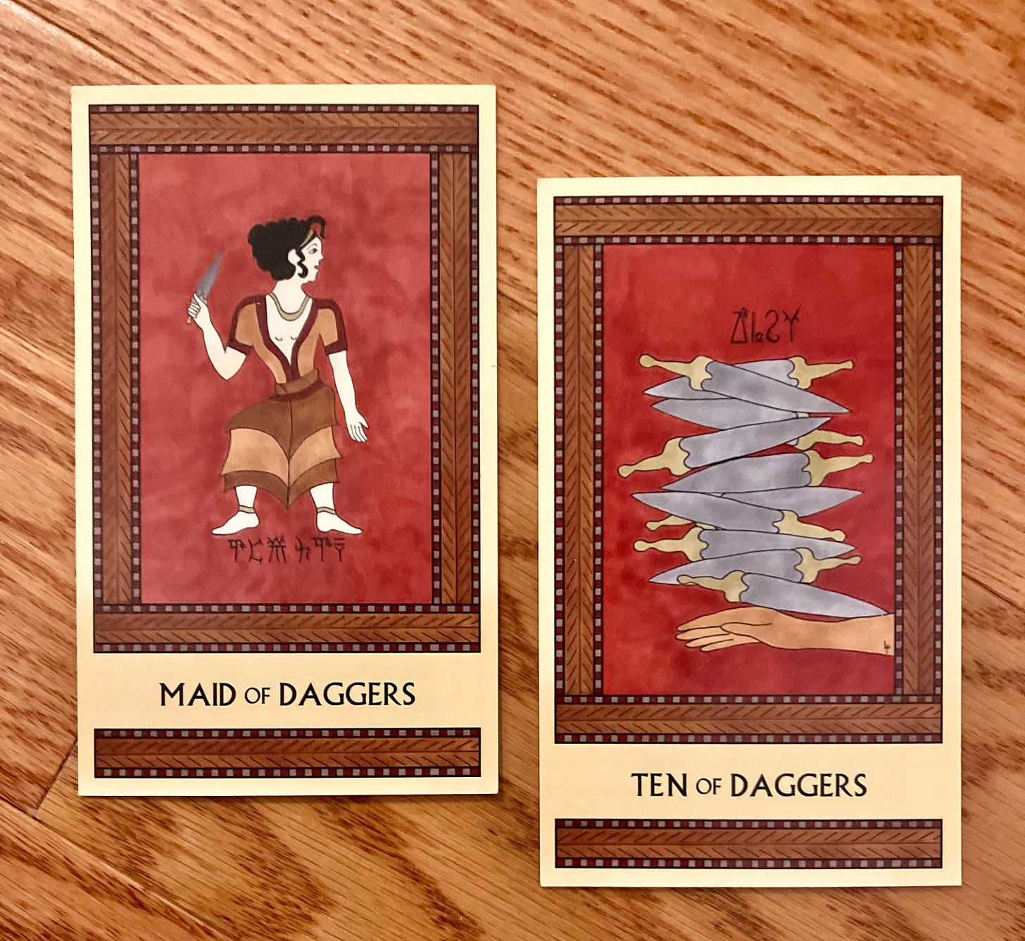 Two Minoan Tarot cards side by side. They're both painted in shades of deep red and brown. The Maid of Daggers shows a Minoan girl standing, her body facing right but her head turned toward the left. She holds up a dagger in her right hand. The Ten of Daggers shows a hand extended out, palm downward, with a stack of ten daggers balanced on top of it.