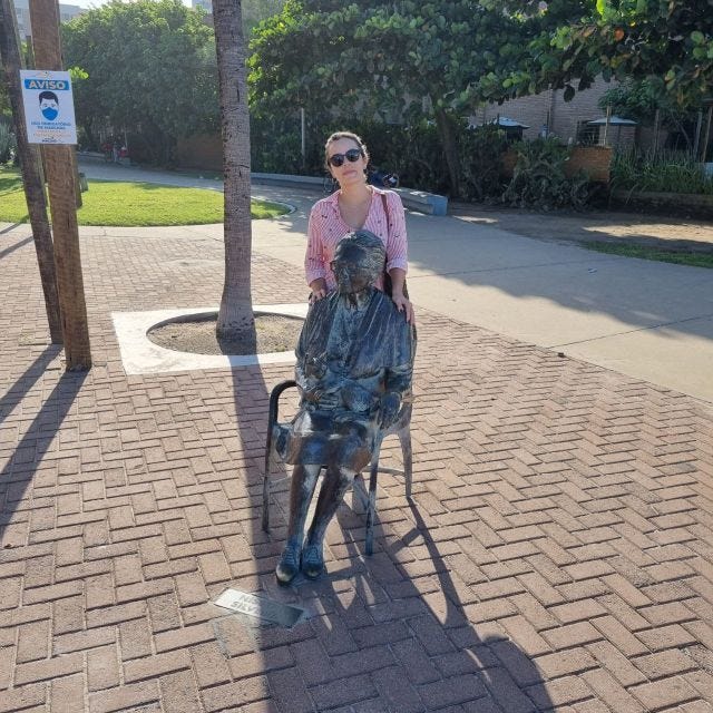 eu e uma estátua de ferro da psiquiatra nise da silveira, na orla de maceió, alagoas. a estátua está sentada numa cadeira, com um gato no colo. estou de pé segurando nos ombros da estátua. o dia está ensolarado e temos sombras na foto.