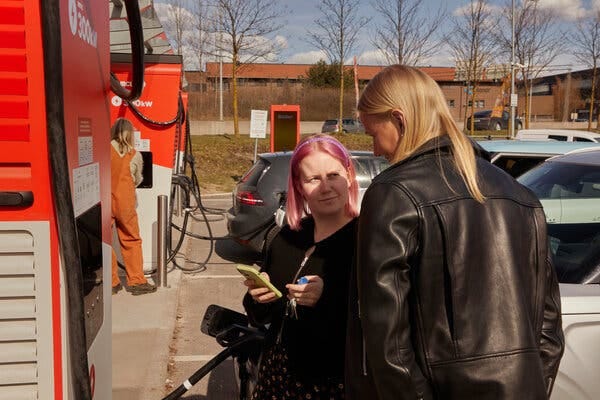 A Circle K executive talking to a customer at a charging station. The executive has pink hair and is holding a phone in her left hand. The customer has blonde hair and is wearing a leather jacket.