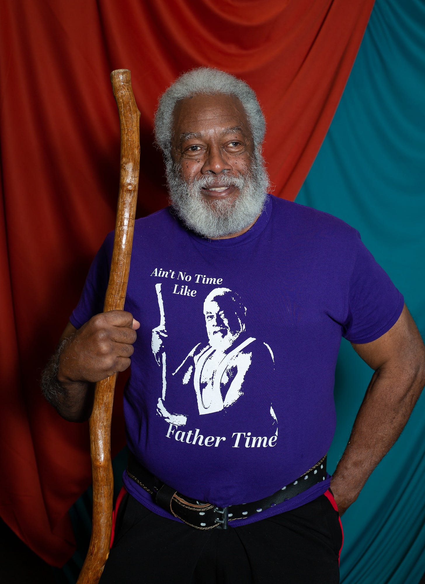 An older man with gray hair and a full white beard smiles confidently with his arms crossed. He wears a purple T-shirt featuring an image of himself holding a staff with the words “Ain’t No Time Like Father Time” printed on it. A draped red and teal fabric backdrop is behind him.